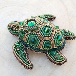 May include: A turtle-shaped brooch crafted with intricate beadwork in shades of emerald and forest green. The shell features faceted green crystals surrounded by metallic gold and green seed beads, creating a textured, dimensional surface.