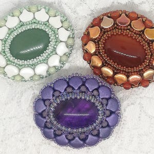 Op de afbeelding: Drie handgemaakte, met kralen bezette broches. Eén heeft een groene ovale steen omringd door witte en groene kralen. Een andere heeft een roodbruine ovale steen met gouden en rode kralen. De derde broche heeft een paarse steen met paarse kralen.