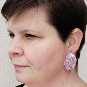 May include: A woman wearing a pair of pink and white beaded oval earrings. The earrings feature a central oval stone, surrounded by detailed beadwork and a scalloped edge. The earrings are a light pink colour.