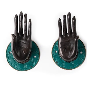 Może przedstawiać: Dwa ozdobne uchwyty do drzwi w kształcie dłoni z uniesionymi palcami. Uchwyty są wykonane z ciemnobrązowego metalu i mają turkusową okrągłą podstawę z kwiatowym wzorem.