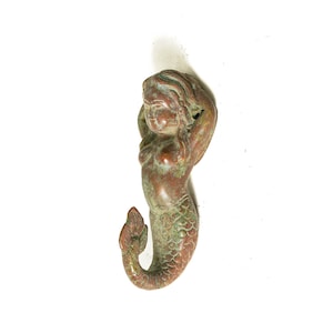 May include: A bronze mermaid figurine with a green patina. The mermaid is depicted with long flowing hair and a tail with scales.