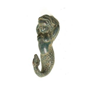 May include: A green mermaid figurine with a detailed tail and a whimsical expression. The mermaid is leaning back with her arms raised above her head.