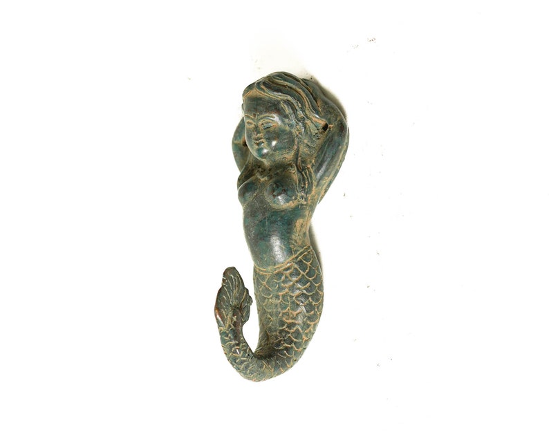 May include: A green mermaid figurine with a detailed tail and a whimsical expression. The mermaid is leaning back with her arms raised above her head.