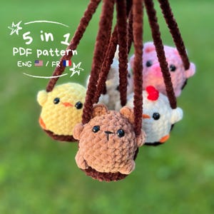 以下が含まれることがあります： 茶色の紐で吊るされた、クマ、ヒヨコ、ブタ、その他2匹を含む、5つの編みぐるみ動物オーナメントのコレクション。オーナメントは様々な色で、黒いビーズの目があります。画像内のテキストは「5 in 1 PDF pattern ENG / FR」と表示されています。