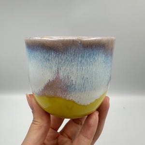 Puede incluir: Una taza de cerámica hecha a mano con un esmalte único. El borde superior es marrón, pasando a azul y blanco, con una base amarilla. La taza se sostiene en una mano, mostrando su forma. La taza probablemente se usa para beber.