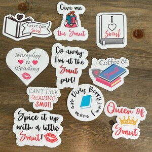 Op de afbeelding: Een set van tien stickers met verschillende boekenthema-ontwerpen en zinnen. De stickers zijn in verschillende kleuren, waaronder roze, rood, blauw en zwart. De zinnen op de stickers zijn: "Give me Smut", "Foreplay is Reading", "Give me the Smut", "I &hearts; Smut", "Go away. I'm at the Smut part!", "Coffee & Smut", "Can't talk, reading smut!", "Spice it up with a little Smut!", "Dirty Books are more fun" en "Queen of Smut".