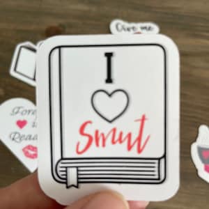 Op de afbeelding: Een witte sticker met een zwarte omtrek van een boek. Het boek heeft een rood hart met het woord "Smut" in rood cursief geschreven. De sticker zegt "Ik hou van Smut" erop.