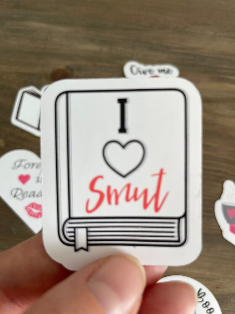 Op de afbeelding: Een witte sticker met een zwarte omtrek van een boek. Het boek heeft een rood hart met het woord "Smut" in rood cursief geschreven. De sticker zegt "Ik hou van Smut" erop.