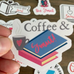 Op de afbeelding: Witte sticker met de tekst "Coffee & Smut!" in zwarte en roze letters. Links op de sticker staat een stomende kop koffie met een hartje. Rechts op de sticker staat een stapel boeken met een blauwe kaft.
