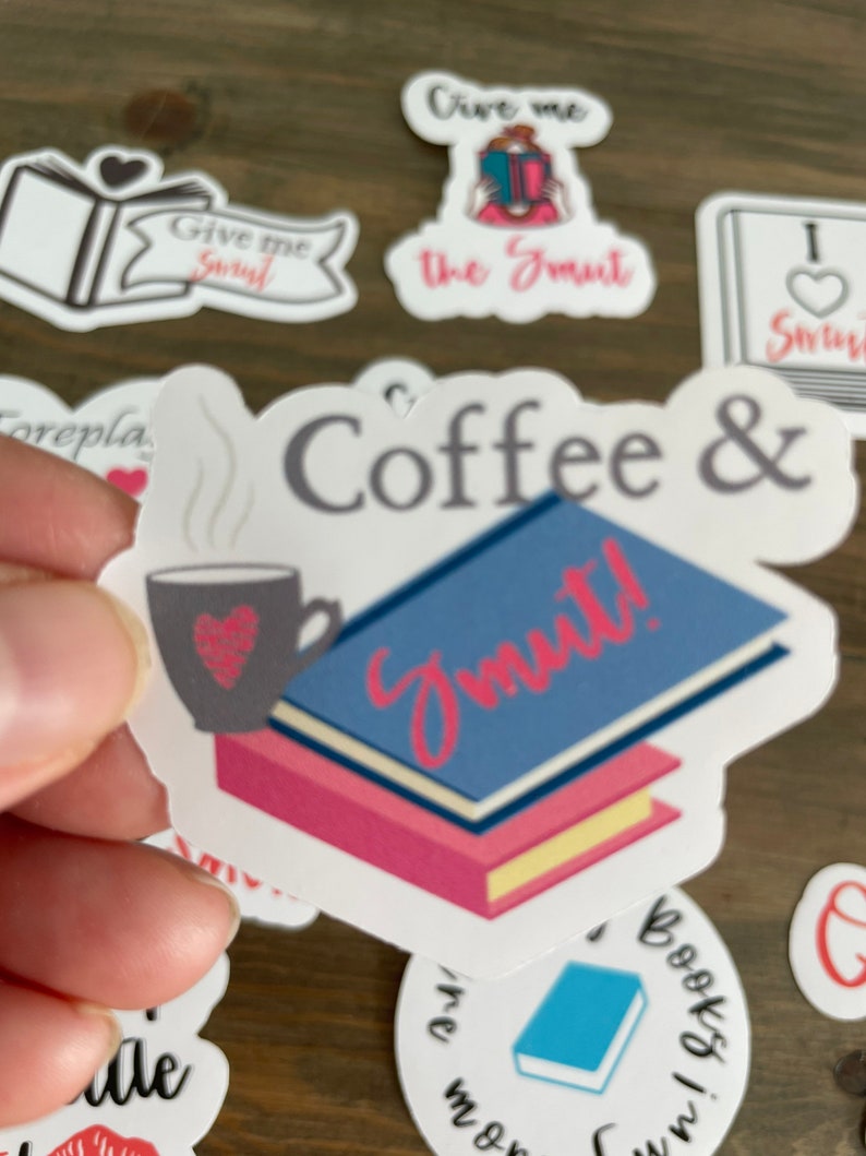 Op de afbeelding: Witte sticker met de tekst "Coffee & Smut!" in zwarte en roze letters. Links op de sticker staat een stomende kop koffie met een hartje. Rechts op de sticker staat een stapel boeken met een blauwe kaft.