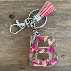 May include: A pink and green resin letter B keyring with dried flower petals. The keyring has a silver chain and a pink tassel.