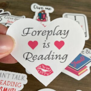 Op de afbeelding: Een witte sticker in de vorm van een hart met de tekst "Foreplay is Reading" in het zwart. Twee roze harten bevinden zich boven het woord "Reading" en een rode lippenstiftkus bevindt zich onder het woord "Reading".