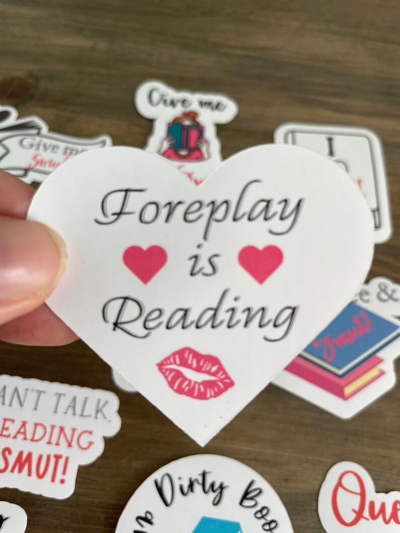 Op de afbeelding: Een witte sticker in de vorm van een hart met de tekst "Foreplay is Reading" in het zwart. Twee roze harten bevinden zich boven het woord "Reading" en een rode lippenstiftkus bevindt zich onder het woord "Reading".