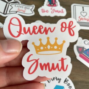 Op de afbeelding: Een witte sticker met de tekst "Queen of Smut" in rode letters. Een gouden kroon zit boven de tekst. De sticker wordt in een hand gehouden.