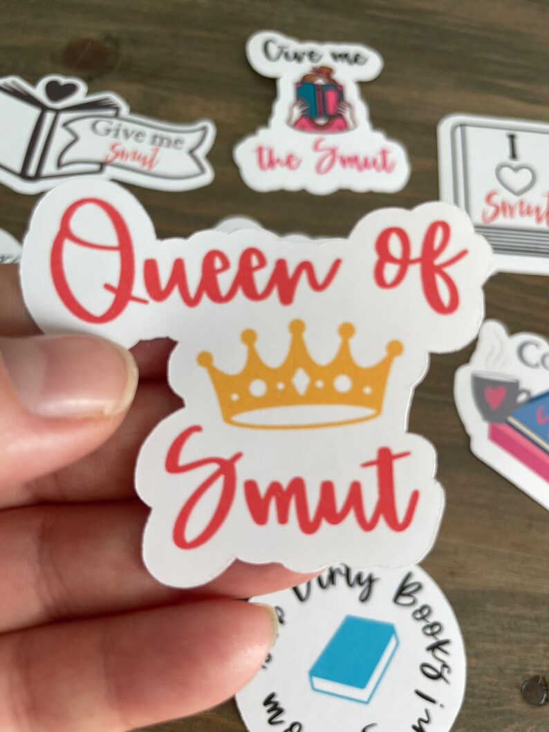 Op de afbeelding: Een witte sticker met de tekst "Queen of Smut" in rode letters. Een gouden kroon zit boven de tekst. De sticker wordt in een hand gehouden.