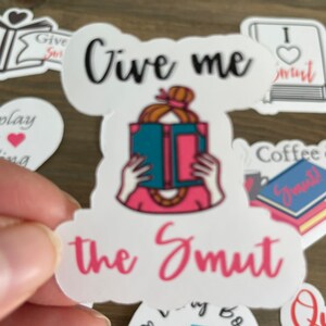 Op de afbeelding: Een witte sticker met een roze en blauwe illustratie van een persoon die een boek leest. De sticker zegt "Give me the Smut" in zwarte en roze letters.