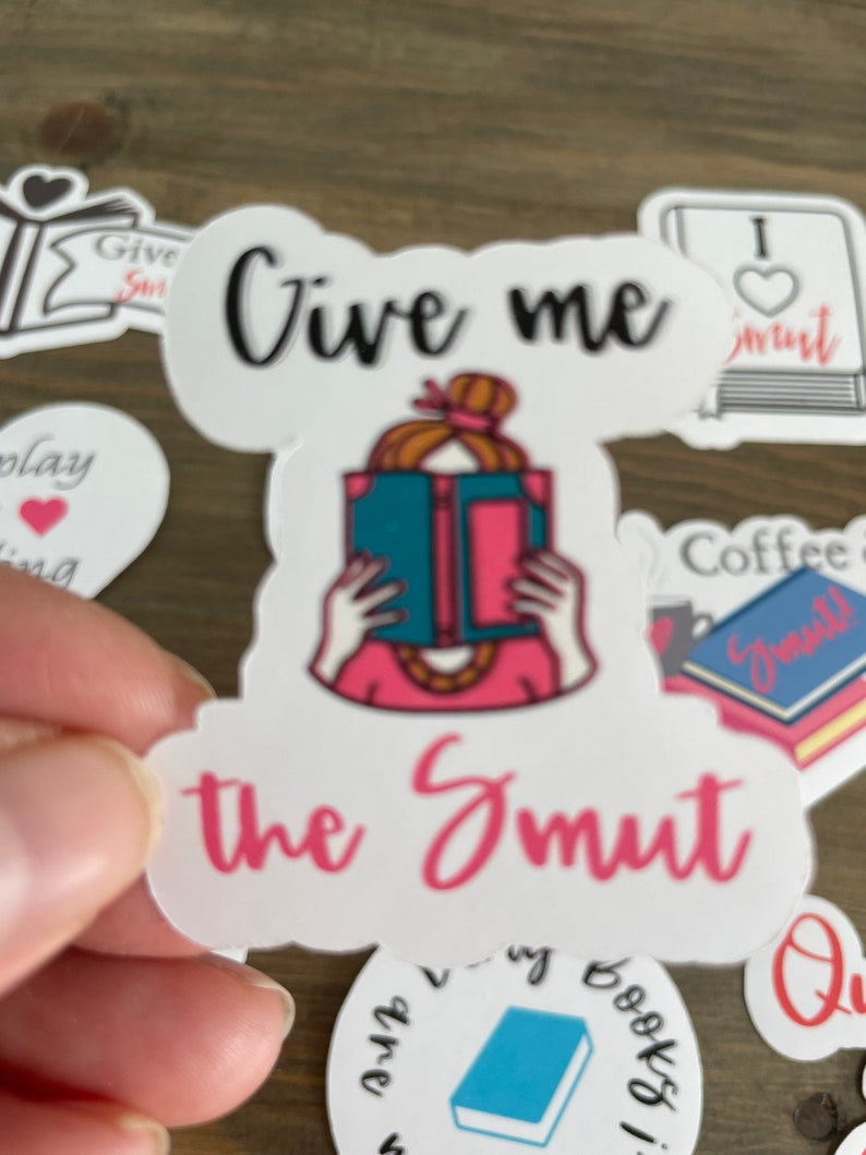 Op de afbeelding: Een witte sticker met een roze en blauwe illustratie van een persoon die een boek leest. De sticker zegt "Give me the Smut" in zwarte en roze letters.