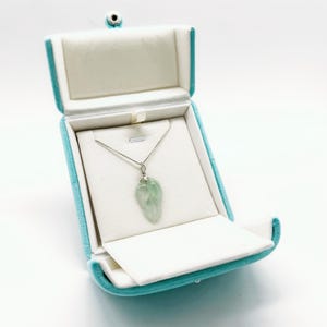 May include: A light green, translucent, leaf-shaped pendant hangs from a thin silver chain. The necklace is displayed inside an open, bright turquoise velvet jewelry box with a white interior lining and a silver-toned snap closure.