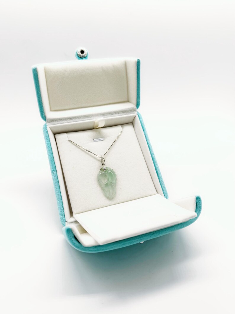 May include: A light green, translucent, leaf-shaped pendant hangs from a thin silver chain. The necklace is displayed inside an open, bright turquoise velvet jewelry box with a white interior lining and a silver-toned snap closure.