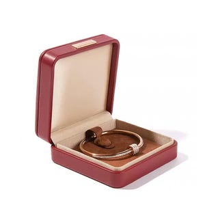 K&ouml;nnte beinhalten: Ge&ouml;ffnete rote Schmuckschatulle mit einem goldenen Armband. Das Armband hat einen diamantbesetzten Abschnitt. Die Schatulle hat einen beigen Innenraum und das Armband liegt auf einem braunen Kissen.