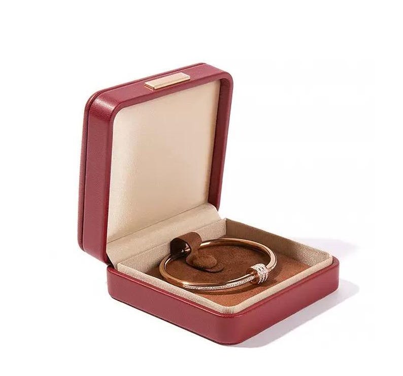 K&ouml;nnte beinhalten: Ge&ouml;ffnete rote Schmuckschatulle mit einem goldenen Armband. Das Armband hat einen diamantbesetzten Abschnitt. Die Schatulle hat einen beigen Innenraum und das Armband liegt auf einem braunen Kissen.