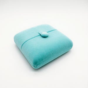 May include: A square, turquoise velvet jewelry box with a button closure. The box is closed and has a soft, plush appearance. The background is plain white.