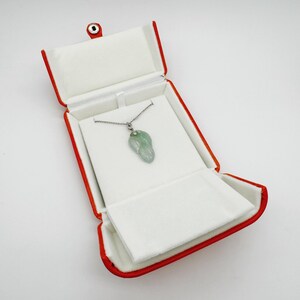 May include: A light green, translucent, leaf-shaped jade pendant hangs from a thin silver-toned chain. The necklace is displayed inside an open, rectangular jewelry box with a vibrant red exterior and a soft, white, padded interior lining.