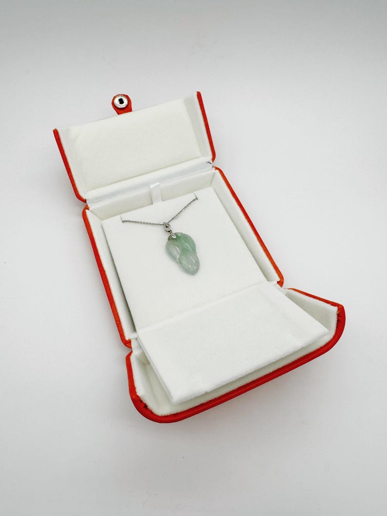 May include: A light green, translucent, leaf-shaped jade pendant hangs from a thin silver-toned chain. The necklace is displayed inside an open, rectangular jewelry box with a vibrant red exterior and a soft, white, padded interior lining.