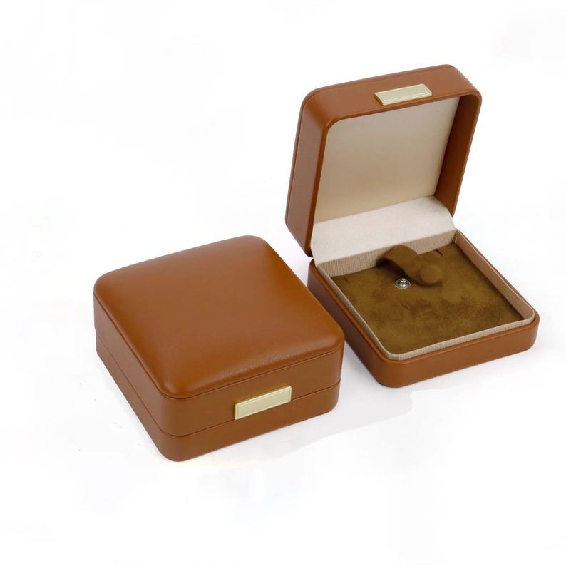 K&ouml;nnte beinhalten: Zwei braune Leder-Schmuckk&auml;stchen, eines offen und eines geschlossen. Die offene Box zeigt ein beiges Innere mit einem dunkelbraunen Samtfutter und einem kleinen Verschluss. Beide Boxen haben einen goldfarbenen Metallakzent.