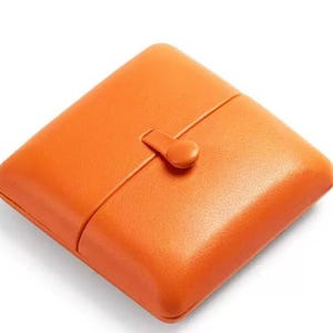 May include: A square, bright orange case with rounded corners and a smooth, textured surface. The case features a central vertical seam and a small, matching orange tab closure positioned in the middle, securing the two halves together.