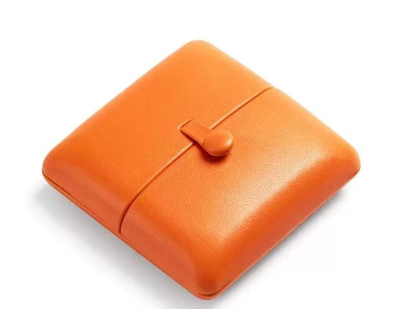 May include: A square, bright orange case with rounded corners and a smooth, textured surface. The case features a central vertical seam and a small, matching orange tab closure positioned in the middle, securing the two halves together.