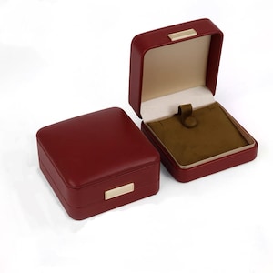 May include: Two burgundy jewelry boxes. One box is closed, and the other is open, revealing a tan interior with a brown velvet lining and a clasp. Both boxes have a gold-colored rectangular accent on the front.