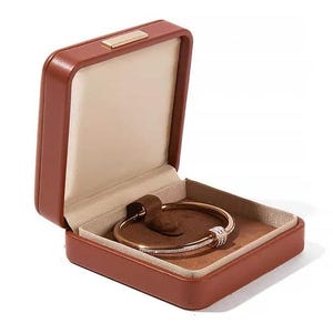 K&ouml;nnte beinhalten: Eine braune Leder-Schmuckschatulle mit einem goldfarbenen Armband darin. Das Armband hat eine Reihe kleiner, klarer Steine und einen dekorativen Verschluss. Die Schatulle ist ge&ouml;ffnet und zeigt ein beiges Innere und einen kleinen, goldfarbenen Verschluss.