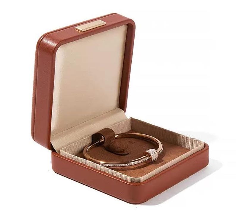 K&ouml;nnte beinhalten: Eine braune Leder-Schmuckschatulle mit einem goldfarbenen Armband darin. Das Armband hat eine Reihe kleiner, klarer Steine und einen dekorativen Verschluss. Die Schatulle ist ge&ouml;ffnet und zeigt ein beiges Innere und einen kleinen, goldfarbenen Verschluss.