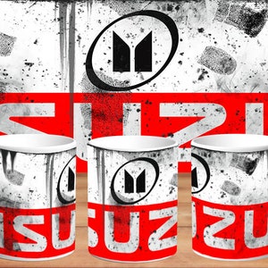 Puede incluir: Taza de cerámica blanca con la palabra "ISUZU" en rojo y un logotipo negro sobre fondo blanco. El diseño incluye salpicaduras negras y un efecto desgastado. La taza se muestra sobre una superficie de madera.