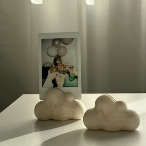 Könnte beinhalten: Zwei weiße wolkenförmige Fotohalter mit einem weißen Polaroid-Foto von Personen, die Ballons halten, im Hintergrund.