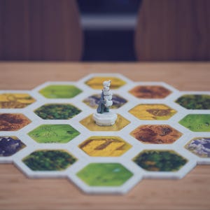 May include: A board game setup featuring a central white figurine placed on a hexagonal tile. The game board is composed of multiple interlocking hexagonal tiles in various colors, including green, yellow, and brown, representing different terrain types.