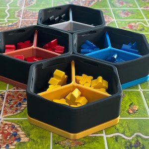 May include: A collection of black, hexagonal game piece organizers with colored dividers. Each container holds wooden game pieces in red, blue, and yellow. The organizers are on a green and brown game board.