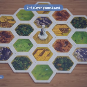 May include: A hexagonal 3-4 player game board featuring various terrain tiles including green fields, brown mountains, and forests. A small white figurine stands on a central tile. Text on the image reads 3-4 player game board, Includes 19 magnetic tile holders, and 19 Squiggle Snaps.