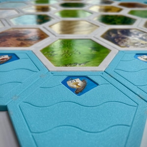 May include: A close-up of a blue hexagonal game board tile with a white border and a small white ship with a number "2:1" on it. The tile is surrounded by other hexagonal tiles.