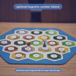 May include: A hexagonal board game frame with a blue border containing multiple hexagonal tiles featuring various terrain illustrations. White circular magnetic number tokens are placed on the tiles. A small red die and a grey figurine are present.