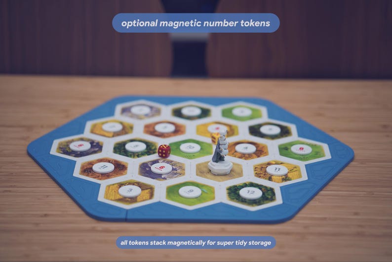 May include: A hexagonal board game frame with a blue border containing multiple hexagonal tiles featuring various terrain illustrations. White circular magnetic number tokens are placed on the tiles. A small red die and a grey figurine are present.
