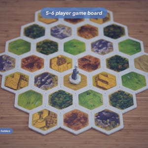May include: A hexagonal 5-6 player game board featuring various terrain tiles including green fields, brown mountains, and yellow wheat fields. A small gray figurine stands in the center. Text on the image reads 5-6 player game board, Includes 30 magnetic land tile holders, and 30 Squiggle Snaps.
