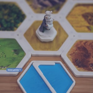 May include: A board game setup featuring hexagonal tiles with landscape illustrations and a white figurine. A blue hexagonal tile with a white S-shaped border is in the foreground. Text reads: and the Squiggle Snap System, loosey goosey, which keeps your tiles securely in place without crimping or squishing your tiles.