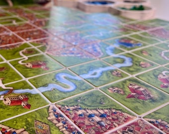 May include: A board game featuring square tiles arranged in a grid, depicting green landscapes, winding blue rivers, and clusters of buildings with red roofs. Small colored game pieces are organized in a white tray in the background.