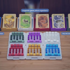 May include: A set of magnetic game piece organizers with six white trays holding small colored tokens in green, purple, white, red, yellow, and blue. Behind them are six colored card trays with lids, each containing a deck of illustrated game cards.