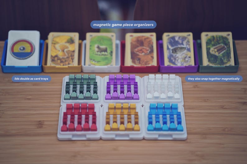 May include: A set of magnetic game piece organizers with six white trays holding small colored tokens in green, purple, white, red, yellow, and blue. Behind them are six colored card trays with lids, each containing a deck of illustrated game cards.
