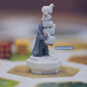 May include: A 3D printed game piece featuring a figure in a grey cloak and green clothing holding a vertical stack of five white sheep. The piece stands on a white circular base. Text on the image reads: from over a dozen other unique Catan upgrades and stack o' sheep robber.