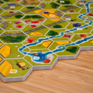 Puede incluir: Un juego de mesa con un mapa modular compuesto por fichas hexagonales. Las fichas muestran campos verdes, parcelas de trigo amarillas, ríos azules serpenteantes y vías de tren grises. Pequeñas ilustraciones de casas y carros están impresas.