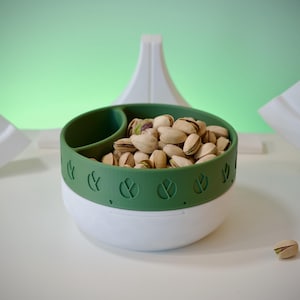 May include: A white and green divided bowl with a lid, filled with pistachios. The bowl has a green band with leaf designs around the rim.